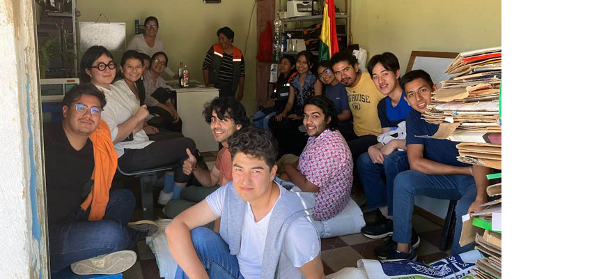 15 people sitting and standing in a cramped and cluttered office.